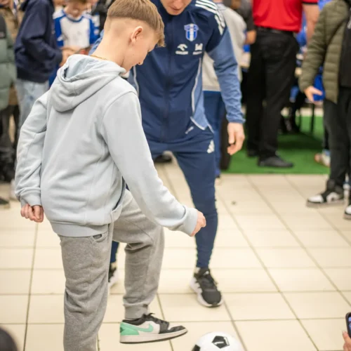 Na zdjęciu widać młodego chłopca grającego w piłkę nożną z dorosłym trenerem w miejscu pełnym dzieci i entuzjastycznej atmosferze.