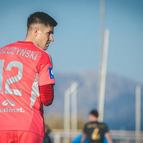 Na zdjęciu widoczny jest piłkarz w różowym stroju sportowym z numerem 12, odwrócony tyłem w kierunku boiska, z górskim krajobrazem w tle.