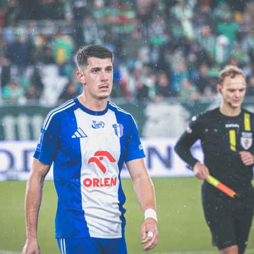 Młody piłkarz w niebiesko-białej koszulce stoi na boisku podczas deszczowego meczu, obok niego sędzia.