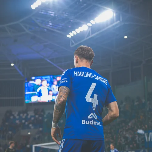 Na zdjęciu widoczny jest piłkarz w niebieskiej koszulce z numerem 4, oglądający mecz na stadionie, przy oświetleniu rozgrywki.