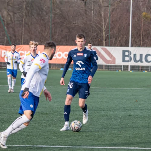 Na zdjęciu widoczna jest akcja meczowa, w której zawodnicy w niebieskich i białych strojach grają na boisku piłkarskim.