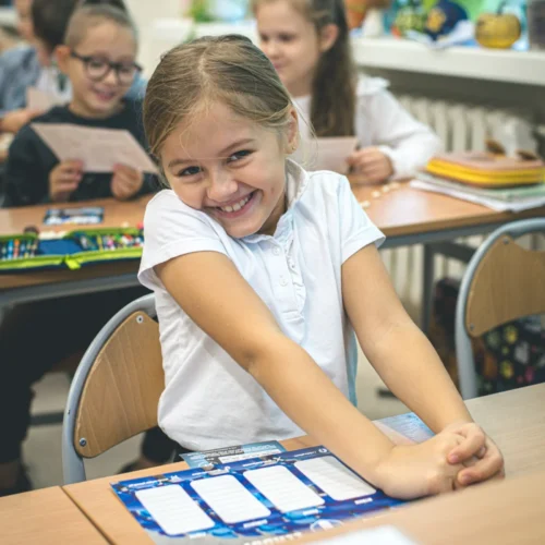 Dziewczynka w koszulce z krótkim rękawem uśmiecha się promiennie, siedząc przy szkolnym biurku w klasie.