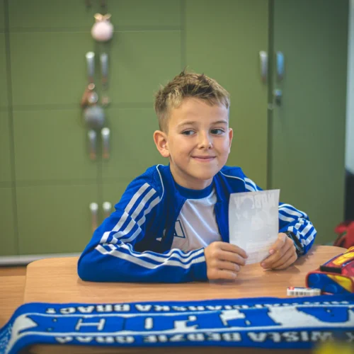 Chłopiec w niebiesko-białym ubraniu siedzi przy biurku w klasie, trzymając kartkę papieru i uśmiechając się do kamery.