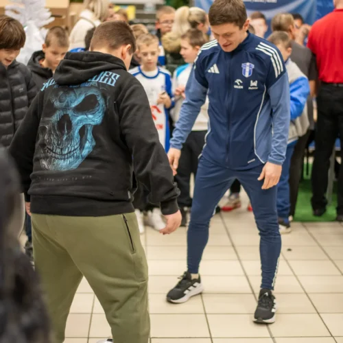 Na zdjęciu widać grupę dzieci otaczających dwóch chłopców, którzy grają w piłkę nożną w sklepie.