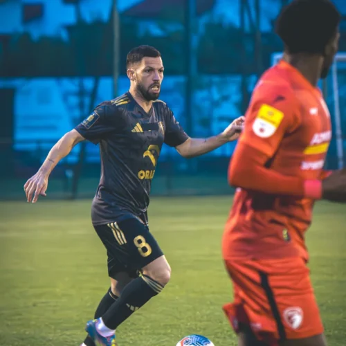 Na zdjęciu znajduje się akcja na boisku piłkarskim, gdzie zawodnik w czarnej koszulce z numerem 8 biegnie z piłką, a w tle widać drugiego gracza w czerwonej koszulce.