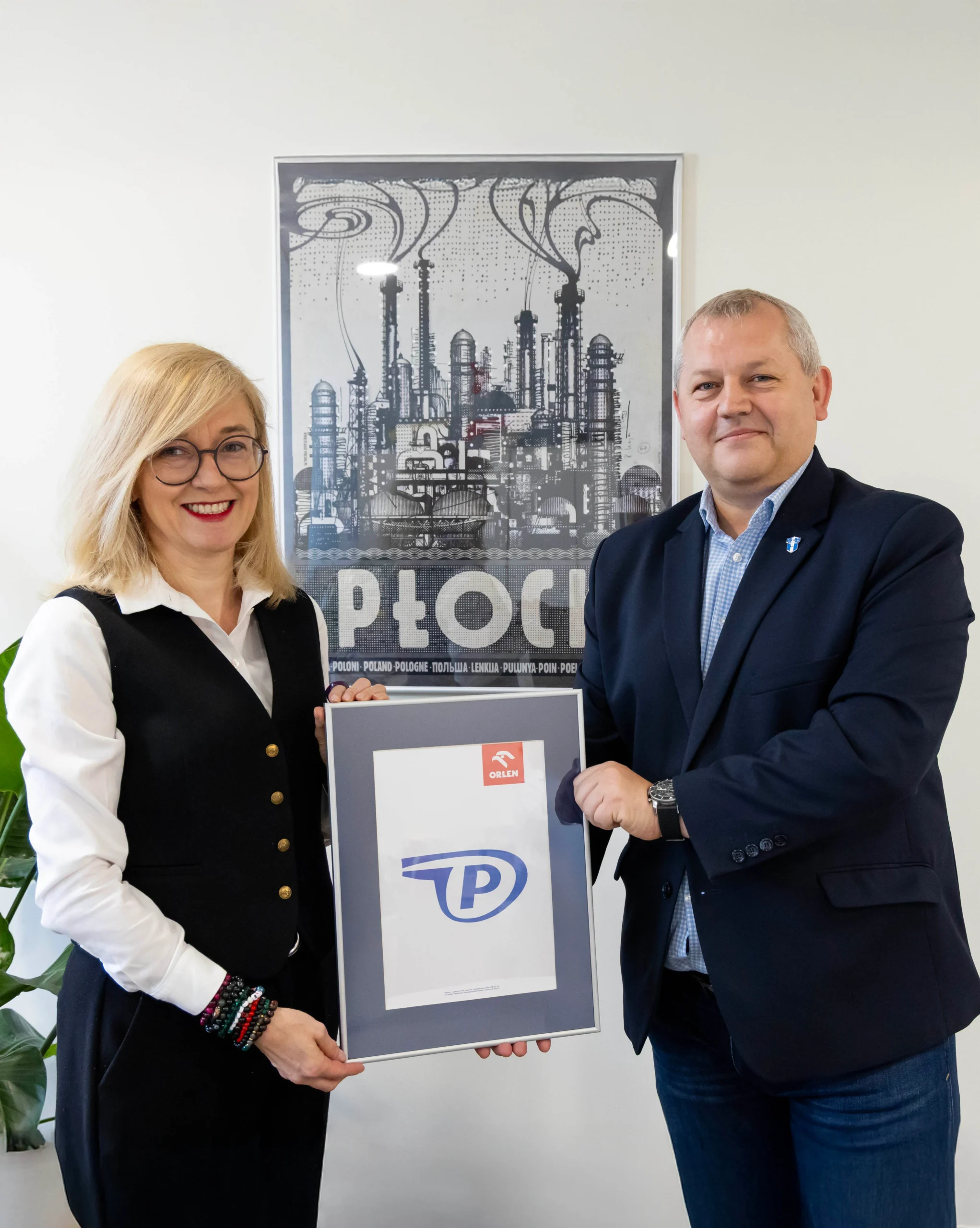 Na zdjęciu dwie osoby trzymają ramkę z logo oraz pozują przed plakatem przedstawiającym przemysł w Płocku.