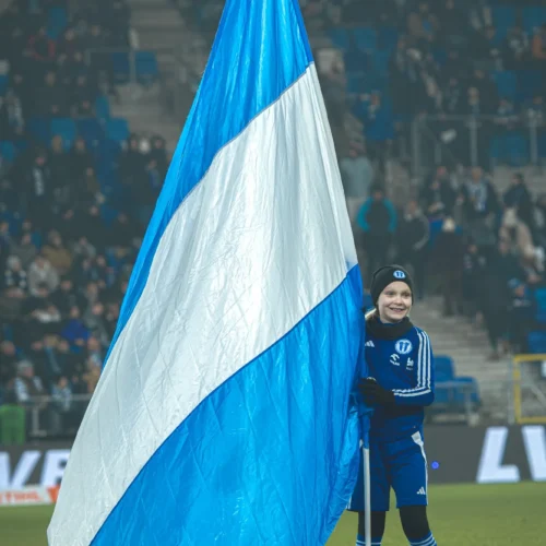 Na zdjęciu widać młodego chłopca trzymającego dużą niebiesko-białą flagę na stadionie w tłumie kibiców.