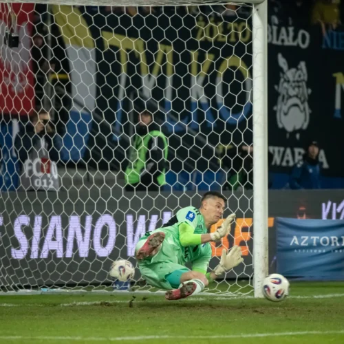 Na zdjęciu widać bramkarza w akcji, który próbuje obronić strzał podczas meczu piłkarskiego, a w tle widoczne są kibice i banery.