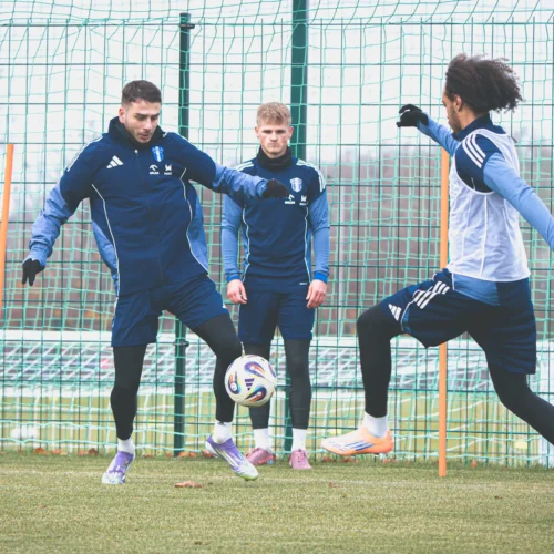 Trzech piłkarzy trenuje na boisku, wykonując ćwiczenia z piłką w towarzystwie siatki.