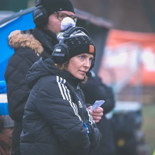 Na zdjęciu widać kobietę w czarnej kurtce i słuchawkach, która obserwuje wydarzenie sportowe, trzymając w ręku notatki.