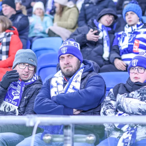 Na zdjęciu widać grupę kibiców siedzących na trybunach stadionu, ubranych w ciepłe kurtki i szaliki w kolorach związanych z ich drużyną.