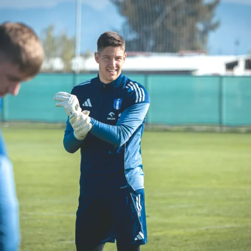 Na zdjęciu widoczny jest bramkarz w niebieskim stroju sportowym, który uśmiecha się i zakłada rękawice, podczas treningu na boisku piłkarskim.