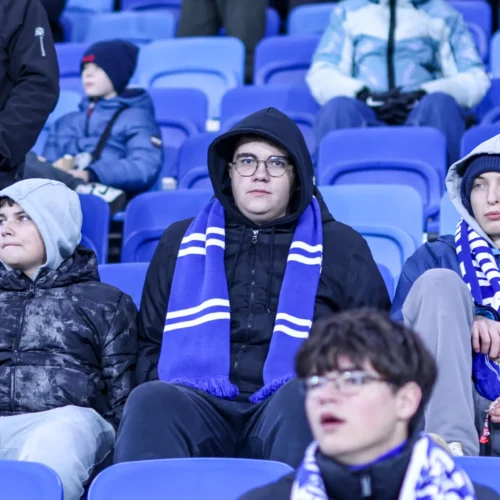 Grupa młodych chłopców ubranych w ciepłe ubrania i szaliki kibicowskie siedzi na trybunach stadionu, obserwując wydarzenie sportowe.