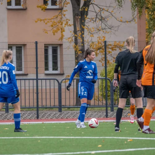 Na zdjęciu widoczne są zawodniczki grające w piłkę nożną, na boisku w strojach w kolorze niebieskim i pomarańczowym, podczas meczu.
