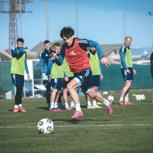 Na zdjęciu widać grupę piłkarzy trenujących na boisku, z jednym zawodnikiem w czerwonym śweterku, który biegnie w kierunku piłki.