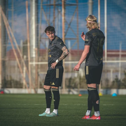 Dwaj zawodnicy w ciemnych strojach piłkarskich rozmawiają na boisku treningowym, położonym na tle nieba i siatki.