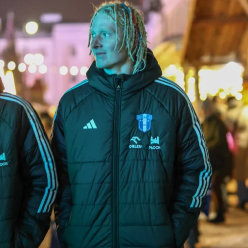 Na zdjęciu widoczny jest mężczyzna w czarnej kurtce zimowej z logotypem, spacerujący w oświetlonej uliczce, obok drugiego mężczyzny.