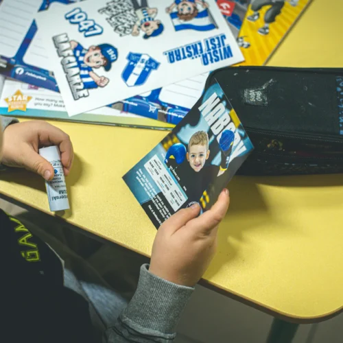 Na zdjęciu widać dziecko trzymające w ręku kartkę z grafiką oraz tubkę kleju na tle biurka z kolorowymi materiałami edukacyjnymi.