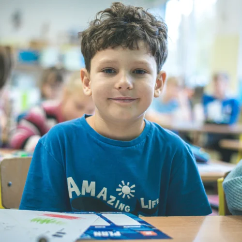 Na zdjęciu widać uśmiechniętego chłopca siedzącego przy biurku w klasie szkolnej, w tle znajdują się inni uczniowie.