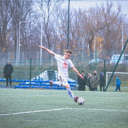 Na zdjęciu znajduje się moment akcji w meczu piłkarskim, gdzie zawodnik w białym stroju wykonuje strzał na bramkę, a bramkarz w zielonym stara się zareagować.