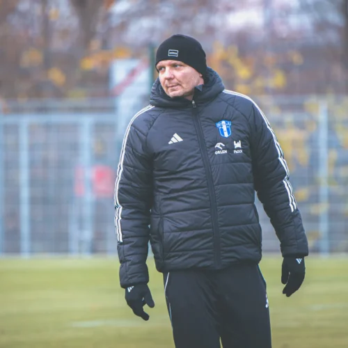 Na zdjęciu widać mężczyznę w czarnej kurtce i czapce, który obserwuje wydarzenia na boisku piłkarskim w chłodny, jesienny dzień.