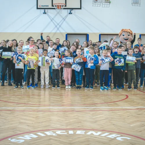 Na zdjęciu znajduje się grupa dzieci oraz dorosłych w gymnasium, wszyscy trzymający nagrody, na tle kosza do koszykówki.