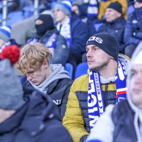Zdjęcie przedstawia grupę kibiców sportowych siedzących na trybunach, w różnych nakryciach głowy i szalikach.