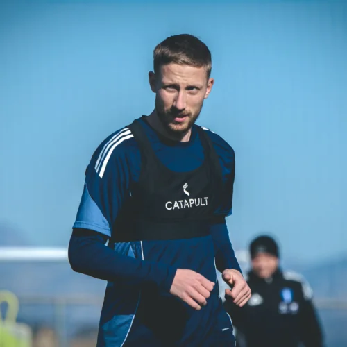 Na zdjęciu widać mężczyznę w sportowym stroju, noszącego czarną kamizelkę z logo, który trenuje na boisku w słoneczną chwilę.