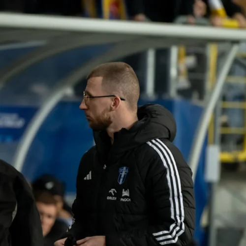 Na zdjęciu widoczny jest mężczyzna w czarnej kurtce z białymi pasami, stojący na stadionie, w otoczeniu innych ludzi.