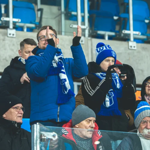 Na zdjęciu widoczni są kibice w zimowych ubraniach, dopingujący swoją drużynę na stadionie, niektórzy trzymający w rękach szaliki.