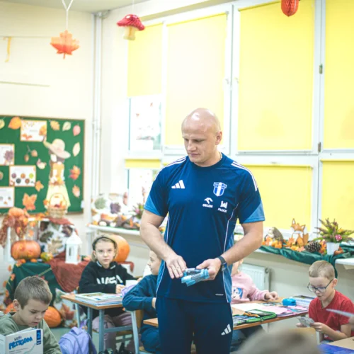 Na zdjęciu widoczna jest klasa, w której nauczyciel w niebieskim stroju sportowym prowadzi zajęcia, a uczniowie siedzą przy biurkach, uczestnicząc w lekcji otoczeni jesiennymi dekoracjami.