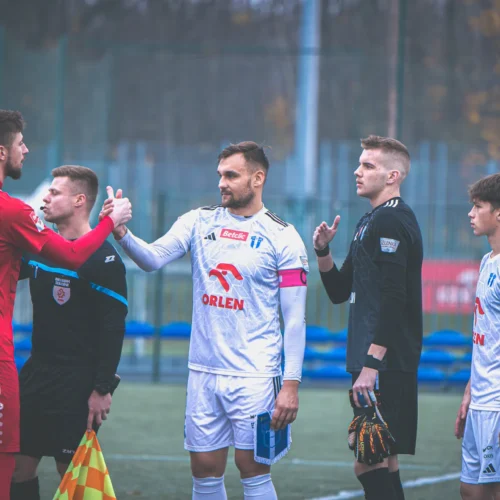 Na zdjęciu widoczni są zawodnicy piłkarscy przygotowujący się do meczu, witający się z sobą na boisku.