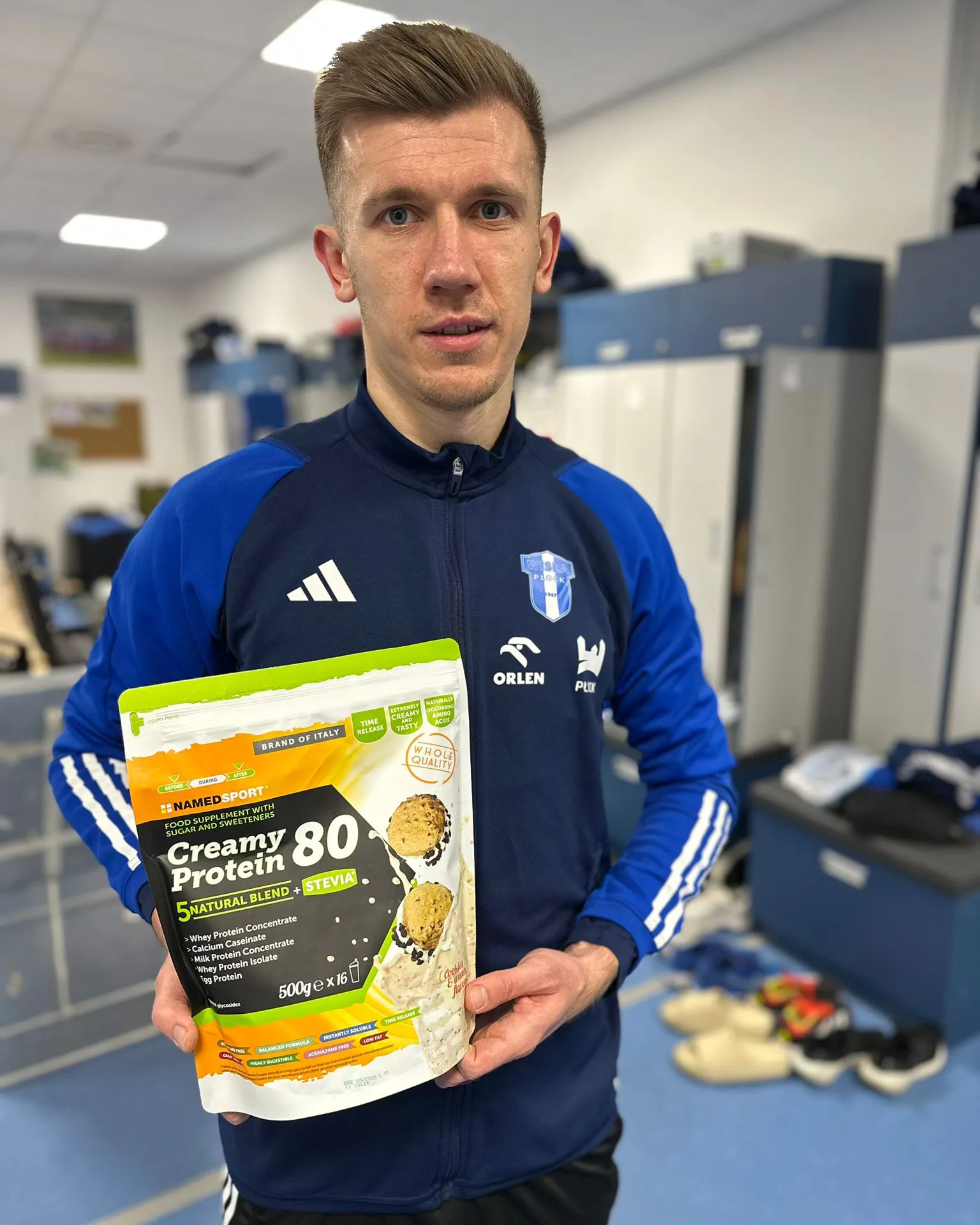 Na zdjęciu widać sportowca trzymającego opakowanie odżywki białkowej Creamy Protein 80 w pomieszczeniu, gdzie znajdują się szafki.