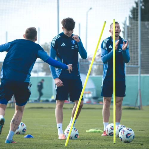 Na zdjęciu trzej piłkarze w niebieskich strojach trenują na boisku, z żółtymi slalomami oraz piłkami w tle.