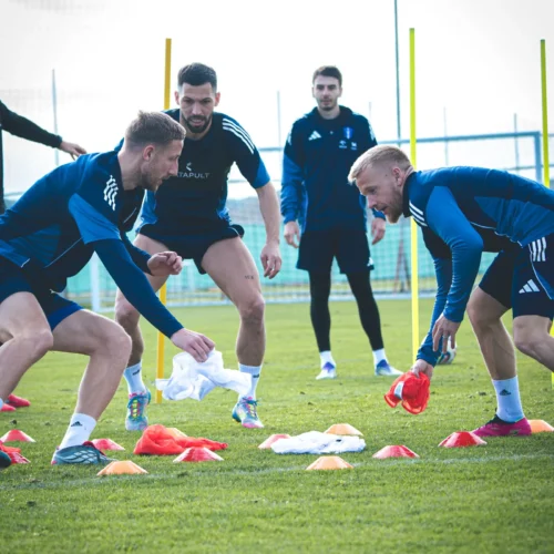 Na zdjęciu widać grupę mężczyzn trenujących na boisku piłkarskim, którzy wykonują ćwiczenia z użyciem pachołków i sprzętu sportowego.