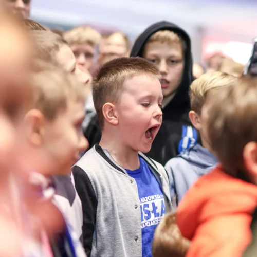 Na zdjęciu widoczna jest grupa dzieci, z których jedno głośno wykrzykuje lub śpiewa, podczas gdy inne przyglądają się z zainteresowaniem.