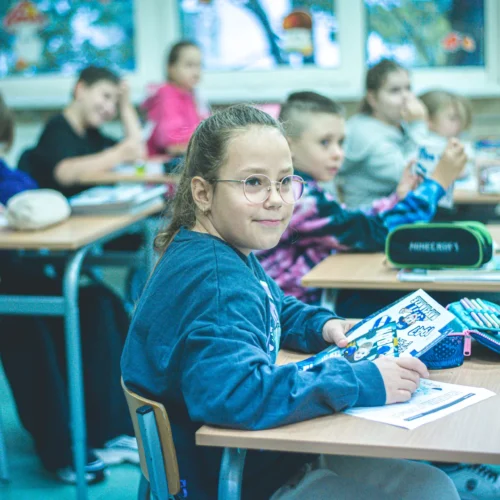 Na zdjęciu widać grupę dzieci w klasie, z których dziewczynka z okularami siedzi na pierwszym planie, trzymając zeszyt i patrząc na kamerę.