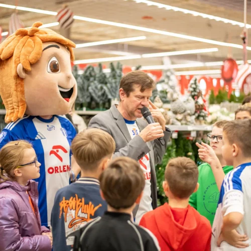 Na zdjęciu widać grupę dzieci słuchających mężczyzny z mikrofonem oraz postać w przebraniu maskotki, podczas wydarzenia w sklepie.