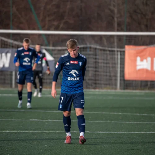 Na boisku piłkarskim młody zawodnik w granatowym stroju przechodzi obok swoich kolegów z drużyny.