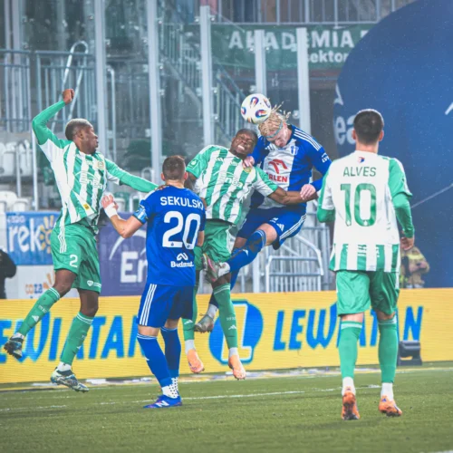 Na zdjęciu widoczni są piłkarze w akcji podczas meczu, walczący o piłkę w deszczowej scenerii.