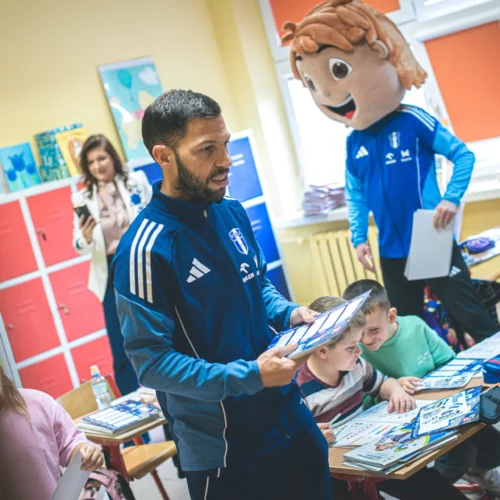 W klasie szkolnej mężczyzna w granatowym dresie trzyma materiały, a w tle widoczna jest postać w dużej głowie maskotki oraz uczniowie przy biurkach.