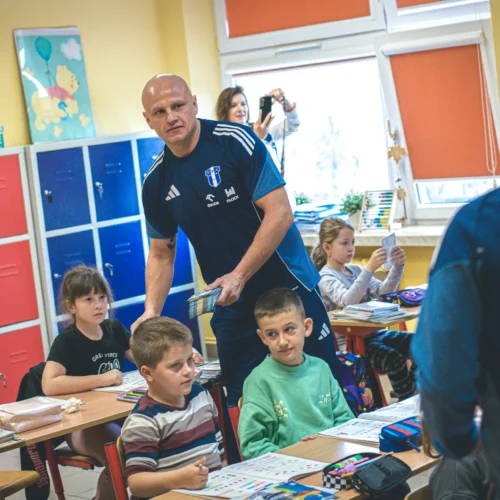 Na zdjęciu widoczna jest klasa szkolna, w której nauczyciel w niebieskim stroju sprzedaje materiały edukacyjne dzieciom siedzącym przy biurkach.