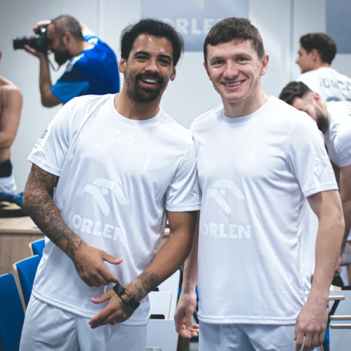 Dwaj sportowcy w białych strojach treningowych uśmiechają się do kamery w tle, podczas gdy inne osoby przygotowują się do zdjęcia.