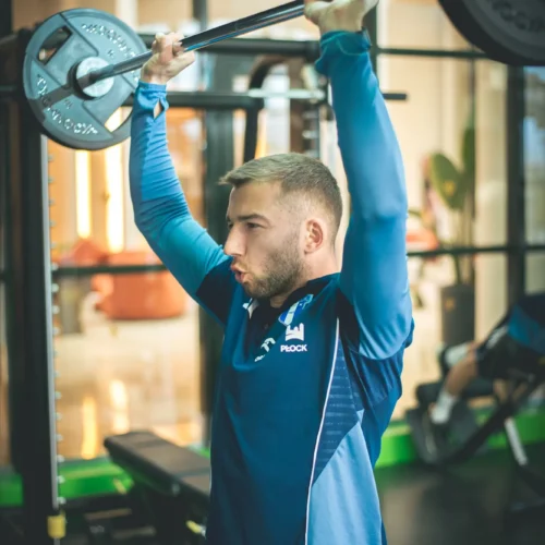 Mężczyzna ćwiczy na siłowni, unosząc sztangę nad głowę w dynamicznej pozie.