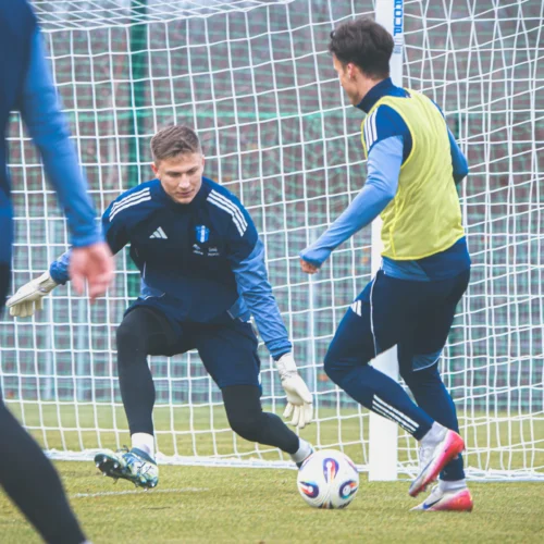 Zdjęcie przedstawia dwóch piłkarzy trenujących na boisku, z jednym z nich w roli bramkarza, próbującego obronić strzał.