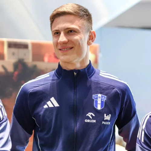 Na zdjęciu widać uśmiechającego się młodego mężczyznę w granatowej bluzie sportowej z logo klubu piłkarskiego.