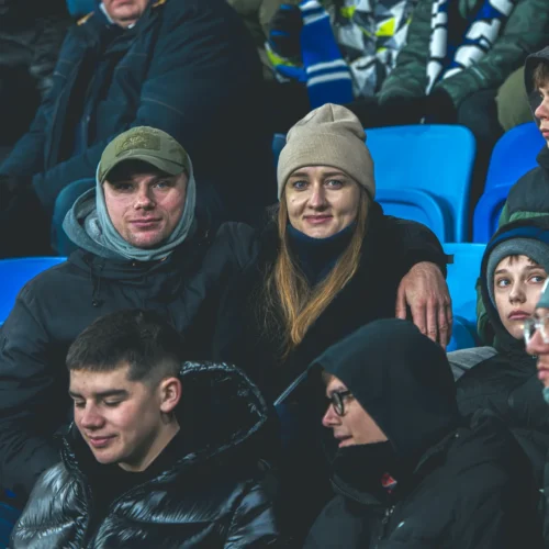 Na zdjęciu widoczna jest grupa kibiców na trybunach, wśród których para siedzi blisko siebie, a w tle widać dodatkowych widzów na stadionie.