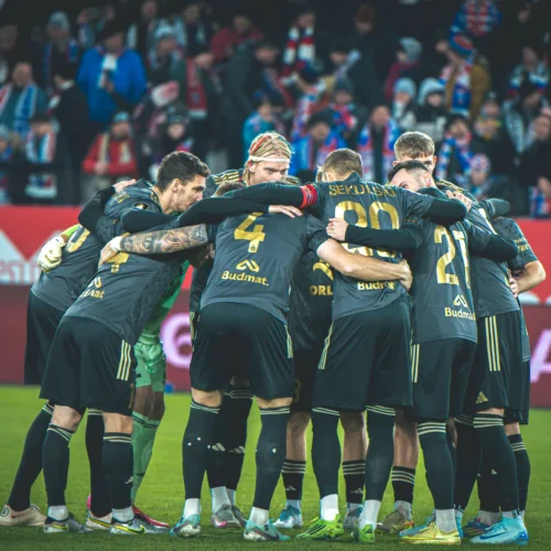 Zespół piłkarski skupiony w okręgu na boisku, przygotowujący się do meczu, z kibicami w tle.