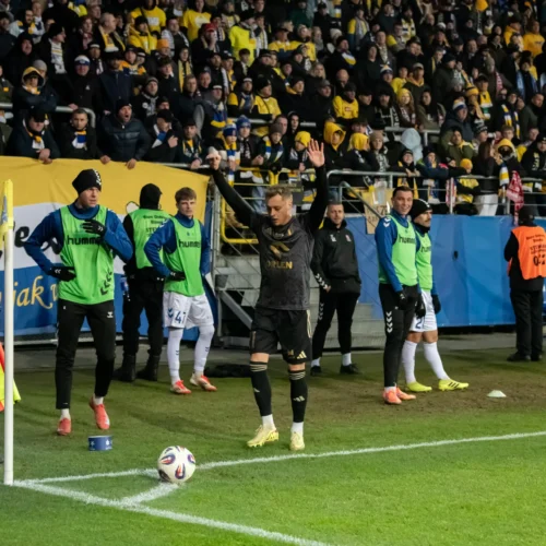 Na zdjęciu widać piłkarza przygotowującego się do wykonania rzutu rożnego, a w tle rozemocjonowani kibice oraz zespół siedzący na ławce rezerwowych.