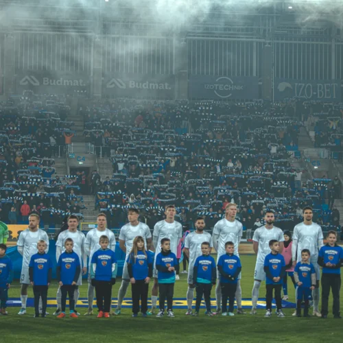 Na zdjęciu widoczni są piłkarze drużyny sportowej stojący na boisku przed meczem, obok dzieci w koszulkach, a w tle widać kibiców z szalikami oraz mgłę unoszącą się nad trybunami.